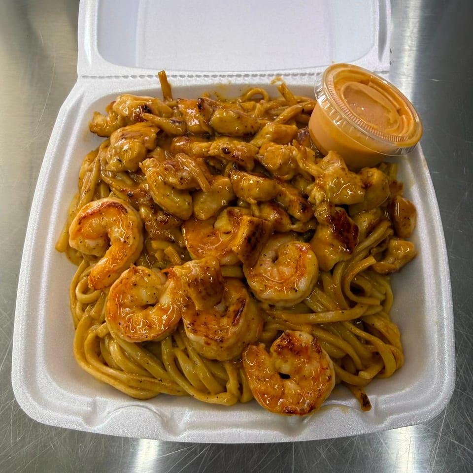 Takeout container filled with grilled shrimp and noodles in creamy sauce, accompanied by a small cup of dipping sauce.