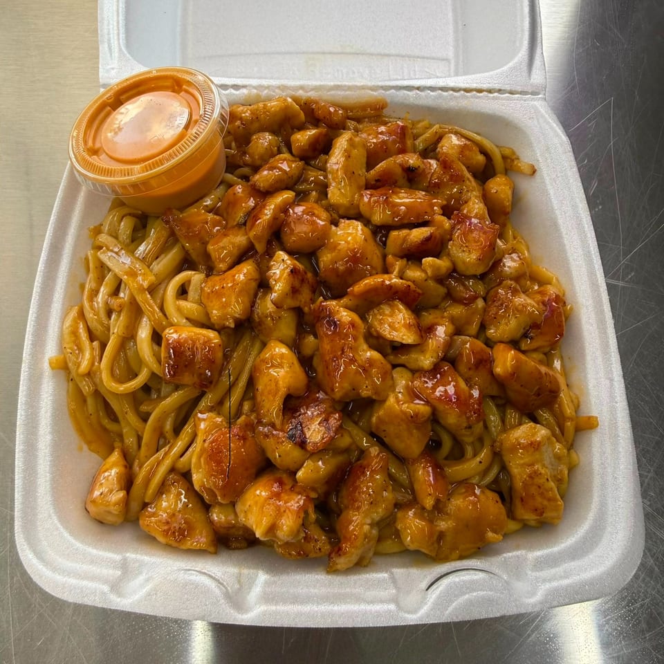 Takeout container filled with glossy sauced chicken, stir-fried noodles, vegetables, and a side of orange dipping sauce.