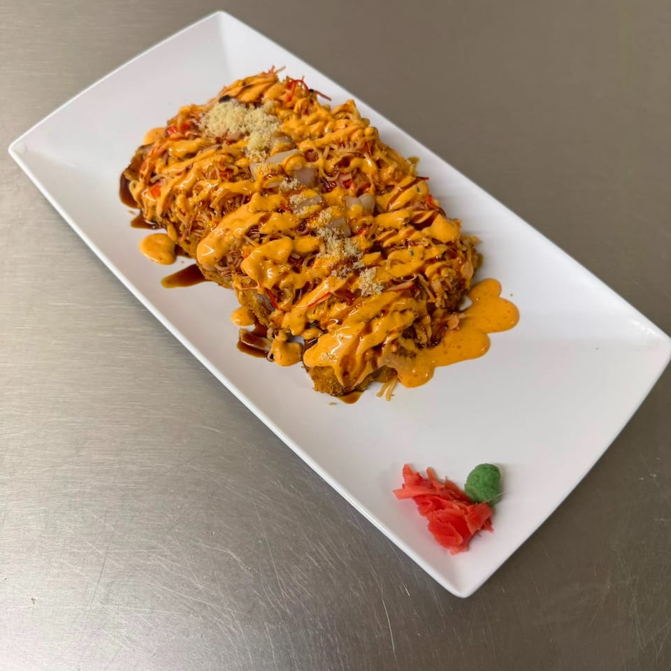 Crispy sushi rolls topped with spicy orange sauce, eel sauce, shredded vegetables, and grated garnish on a white rectangular plate.